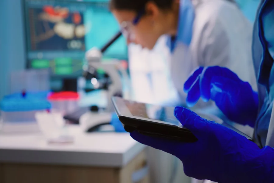 Foto aus dem Labor - Forscher bedient Tablet während Forscherin im Hintergrund in ein Mikroskop blickt