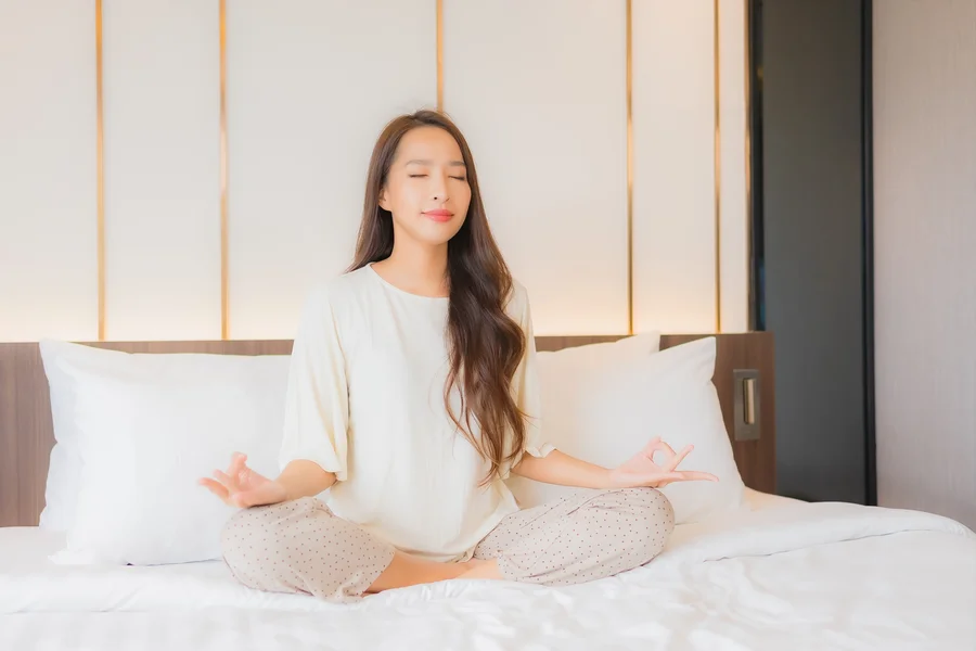 Frau mit langen braunen Haaren sitzt in weißem Gewand auf Bett und meditiert